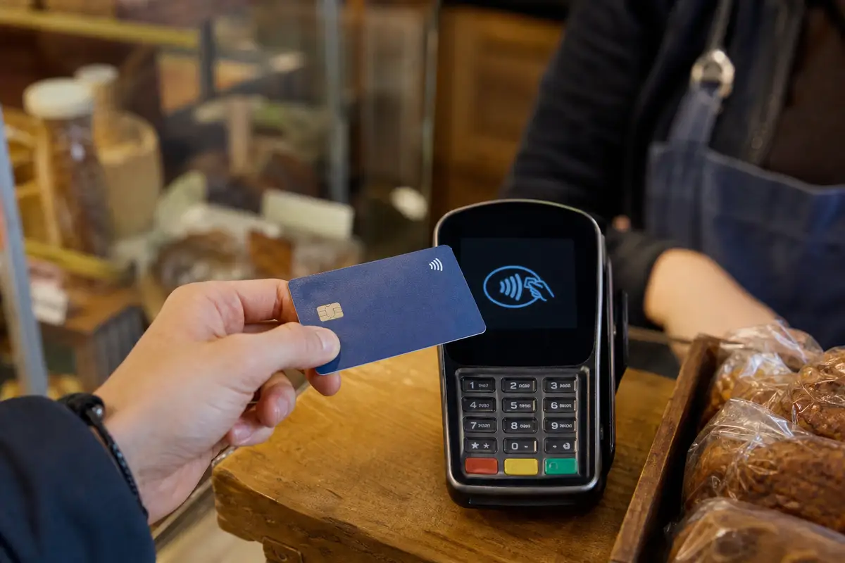 Main tenant une carte bancaire près d’un terminal de paiement sans contact dans un commerce, écran du terminal visible