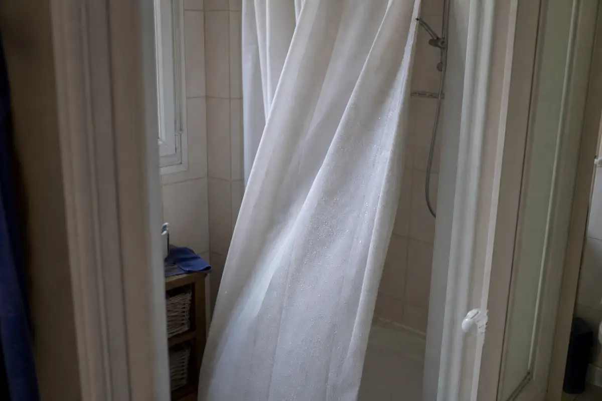 Rideau de douche blanc légèrement aspiré vers l’intérieur d’une cabine, gouttes d’eau visibles sur le tissu